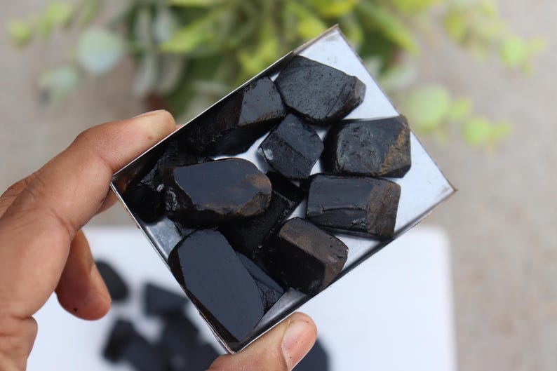 May include: A collection of raw, black tourmaline stones displayed in a small, square metal tray. The stones have a rough, natural texture and varying shapes. The tray is held in a hand, with a blurred background.