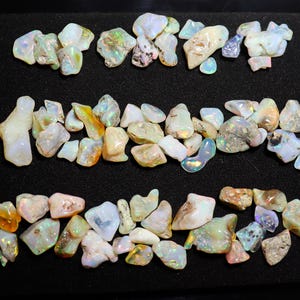 May include: A collection of white, iridescent opal stones of various sizes and shapes. The stones are arranged in three rows on a black background.