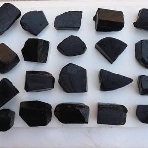 May include: An overhead shot displays a collection of rough, black tourmaline stones. The stones vary in size and shape, with some appearing rectangular and others more organic. They are arranged on a white surface, showcasing their dark, opaque texture.