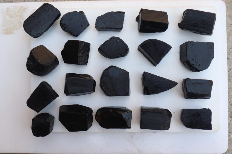 May include: An overhead shot displays a collection of rough, black tourmaline stones. The stones vary in size and shape, with some appearing rectangular and others more organic. They are arranged on a white surface, showcasing their dark, opaque texture.