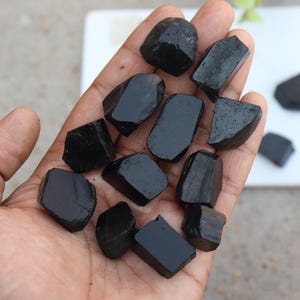 May include: A hand holds a collection of polished, black tourmaline stones. The stones vary in size and shape, with a glossy, reflective surface. The background is a neutral white surface, highlighting the dark stones. These stones are often used for their metaphysical properties.