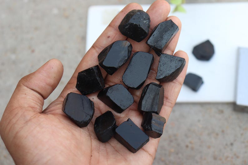 May include: A hand holds a collection of polished, black tourmaline stones. The stones vary in size and shape, with a glossy, reflective surface. The background is a neutral white surface, highlighting the dark stones. These stones are often used for their metaphysical properties.