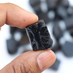 May include: A close-up of a black, rough-textured gemstone held between fingers. The stone has a glossy surface and irregular edges. Other similar stones are blurred in the background.