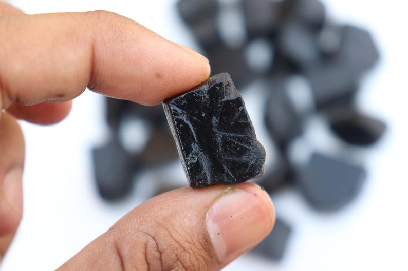 May include: A close-up of a black, rough-textured gemstone held between fingers. The stone has a glossy surface and irregular edges. Other similar stones are blurred in the background.