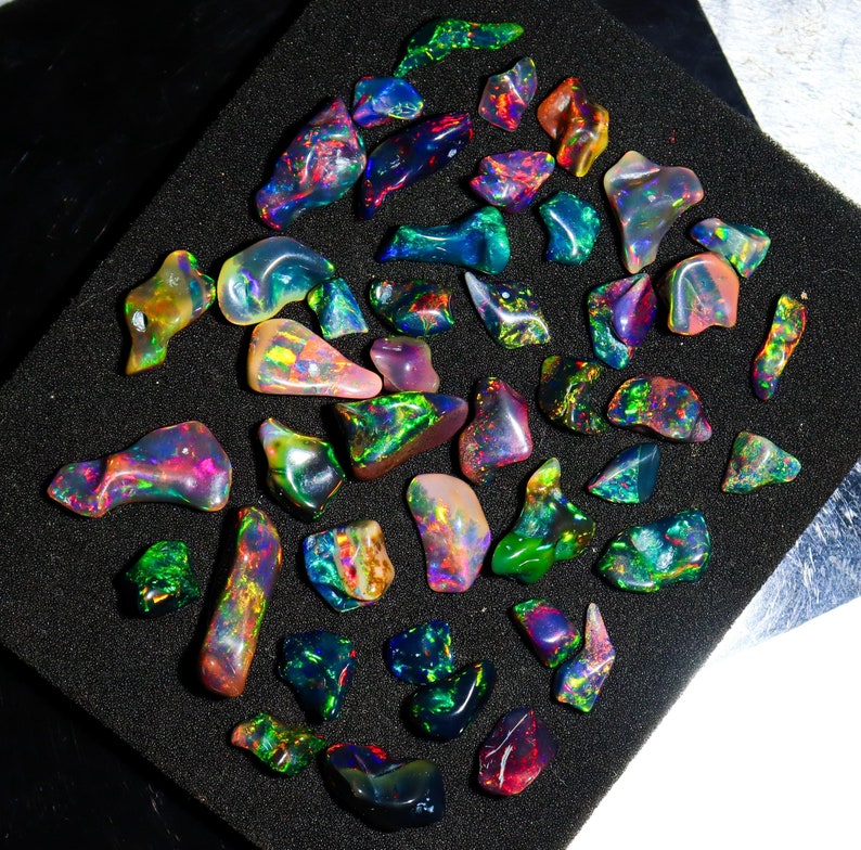 May include: A collection of polished opal gemstones in various shapes and sizes. The opals display a vibrant play of color, including blues, greens, reds, and yellows. They are arranged on a black, textured surface, showcasing their iridescent qualities.