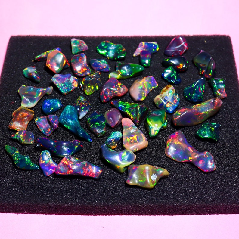 May include: A collection of iridescent opal gemstones in various shapes and sizes. The opals display a vibrant play of color, including green, blue, red, and yellow hues. They are arranged on a black surface against a pink background.
