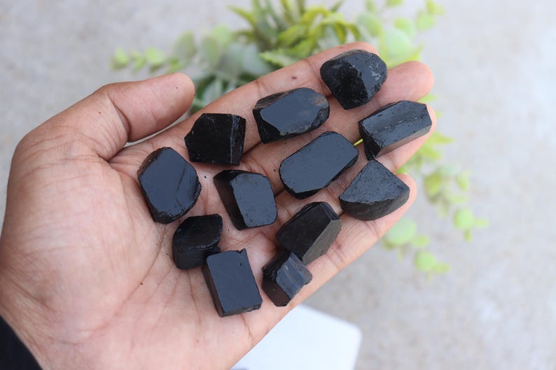 May include: A hand holding several rough, black tourmaline stones. The stones vary in size and shape, with a dark, glossy appearance. The background is blurred, with hints of green foliage.