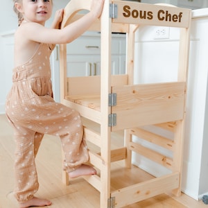May include: A young child stands on a wooden learning tower with the words "Sous Chef" printed on the top. The tower has three steps and is made of natural wood.