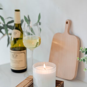 May include: A white candle with a ribbed glass jar is lit and sitting on a wooden coaster. A glass of white wine and a bottle of wine are in the background.