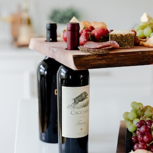 May include: A wooden serving tray with two bottles of red wine, one with the label "Cacciata 2018" and the other with a black label. The tray is filled with cheese, salami, grapes, and strawberries.