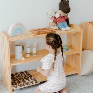 May include: A young girl plays with wooden alphabet blocks and a rainbow toy on a wooden shelf. The shelf has two shelves and is made of natural wood. The girl is wearing a pink and white striped dress and has her hair in two braids.