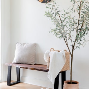 May include: A wooden bench with black metal legs, a white throw blanket in a woven basket, and a potted olive tree. A round painting of flowers hangs on the wall behind the bench.
