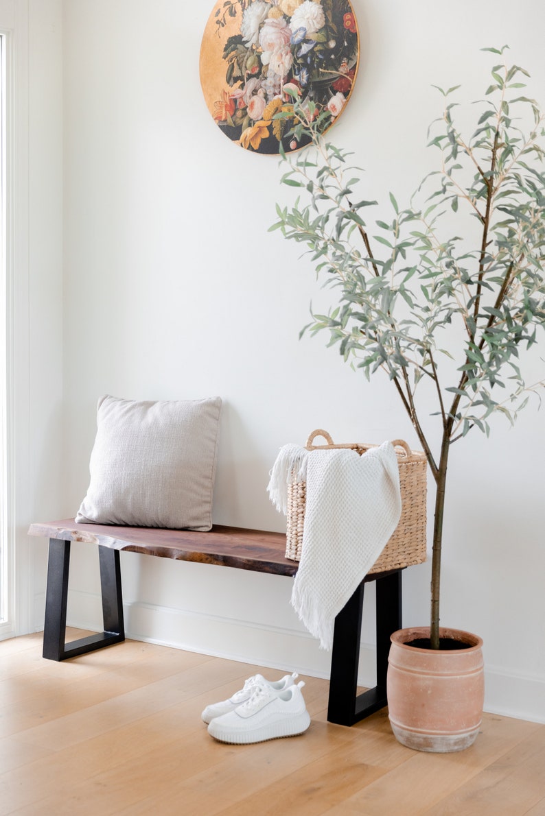 May include: A wooden bench with black metal legs, a white throw blanket in a woven basket, and a potted olive tree. A round painting of flowers hangs on the wall behind the bench.