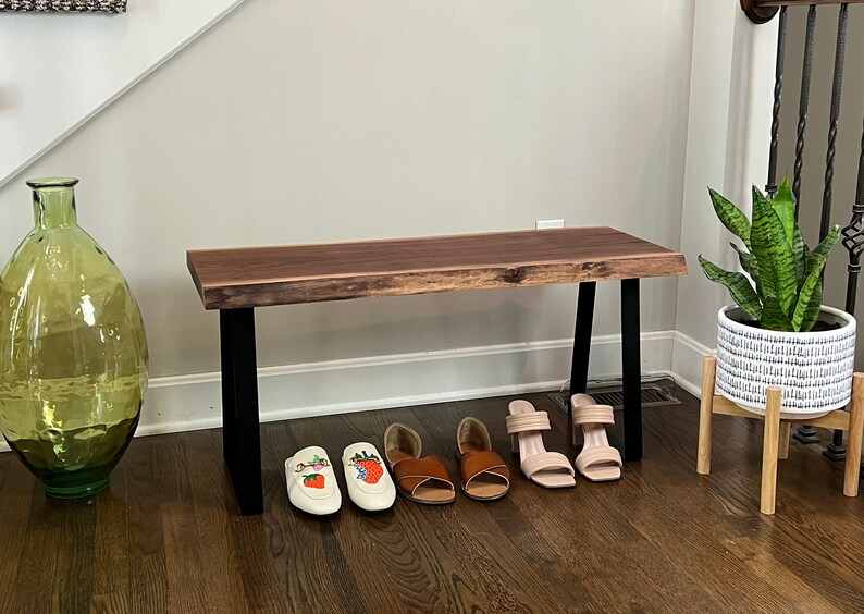 May include: A wooden bench with a live edge top and black metal legs. The bench is in a hallway and has shoes and a plant on the floor in front of it.