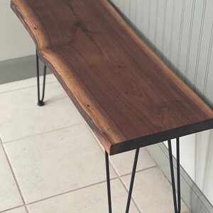 May include: A wooden bench with a live edge top and black hairpin legs. The bench is made of walnut wood and has a natural finish.
