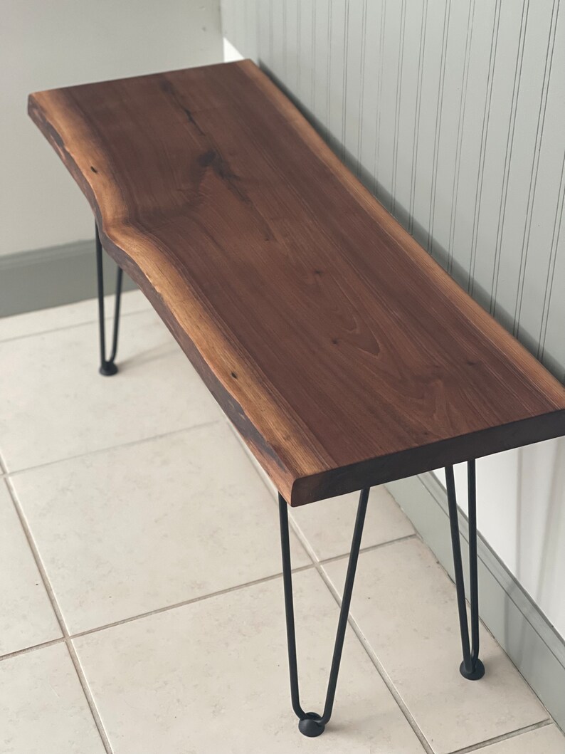 May include: A wooden bench with a live edge top and black hairpin legs. The bench is made of walnut wood and has a natural finish.