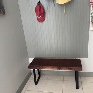 May include: A dark brown wooden bench with black metal legs. The bench is in a hallway with a light gray wall and white tile floor.