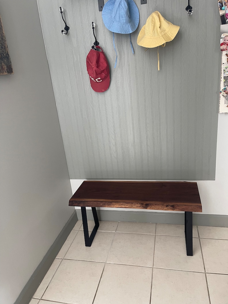 May include: A dark brown wooden bench with black metal legs. The bench is in a hallway with a light gray wall and white tile floor.