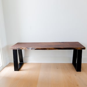 May include: A wooden bench with a live edge top and black metal legs. The bench is in a room with white walls and hardwood floors.
