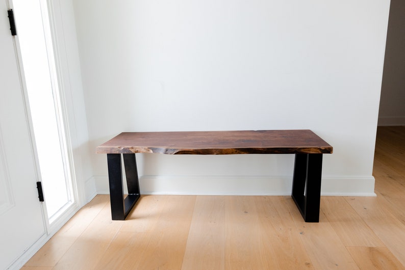 May include: A wooden bench with a live edge top and black metal legs. The bench is in a room with white walls and hardwood floors.