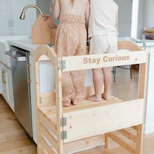 May include: Two children stand on a wooden learning tower with the words "Stay Curious" engraved on the top. The tower has two steps and is made of natural wood.