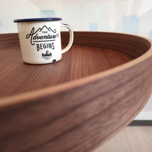 K&ouml;nnte beinhalten: Eine wei&szlig;e Emailletasse mit blauem Rand und einer schwarzen Grafik, auf der "The Adventure Begins" mit einem kleinen Kanu-Bild steht. Die Tasse steht auf einem runden Holztablett.