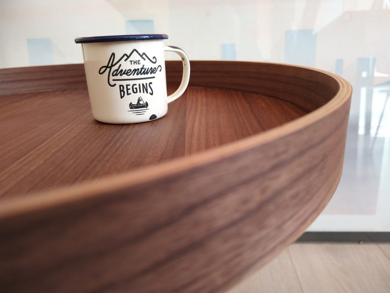 K&ouml;nnte beinhalten: Eine wei&szlig;e Emailletasse mit blauem Rand und einer schwarzen Grafik, auf der "The Adventure Begins" mit einem kleinen Kanu-Bild steht. Die Tasse steht auf einem runden Holztablett.