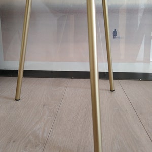 K&ouml;nnte beinhalten: Ein goldfarbener Metalltischst&auml;nder mit drei Beinen. Der St&auml;nder hat eine runde Oberseite und steht auf einem hellen Holzboden.