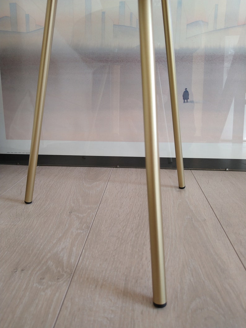 K&ouml;nnte beinhalten: Ein goldfarbener Metalltischst&auml;nder mit drei Beinen. Der St&auml;nder hat eine runde Oberseite und steht auf einem hellen Holzboden.