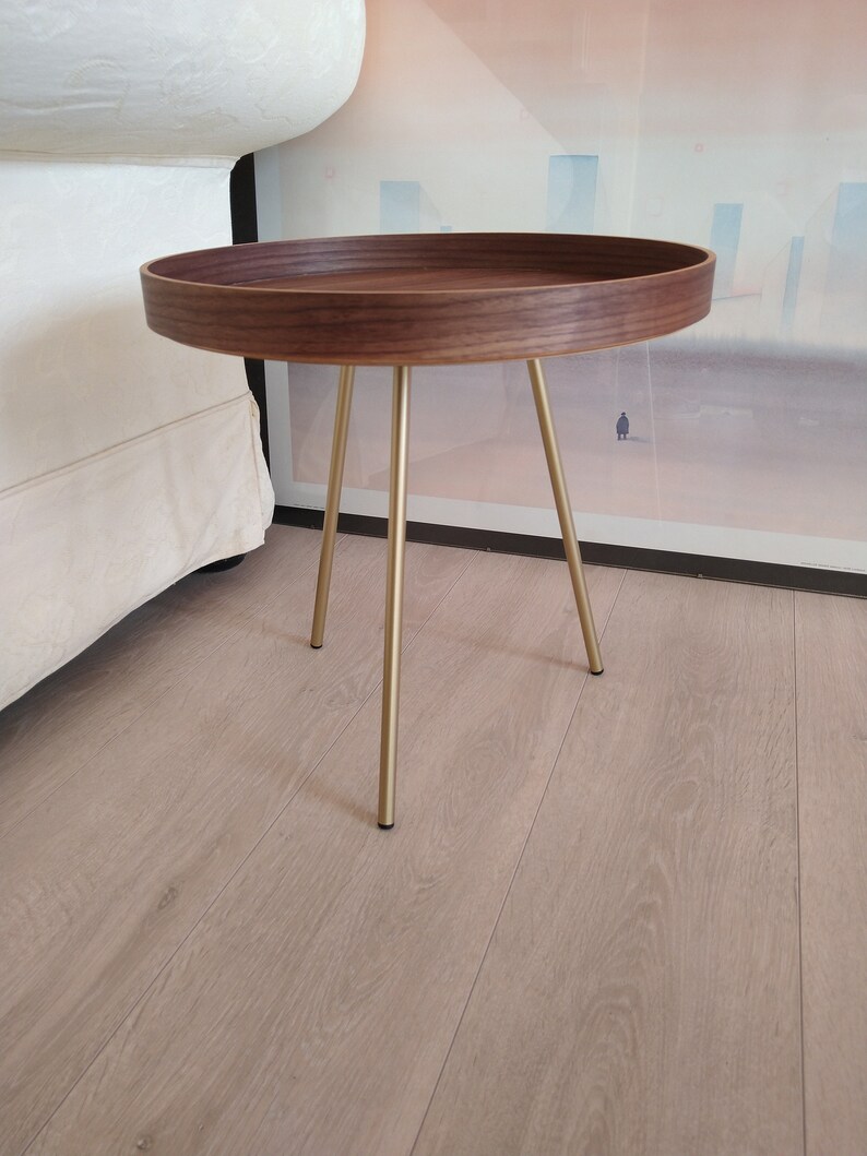 K&ouml;nnte beinhalten: Ein runder Holz-Beistelltisch mit Walnuss-Finish und einem goldenen Metallgestell. Der Tisch hat drei Beine und einen erh&ouml;hten Rand um die Oberseite.