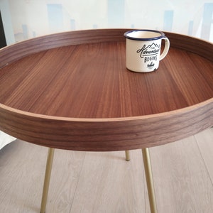 K&ouml;nnte beinhalten: Ein runder Holz-Beistelltisch mit einem goldenen Metallgestell. Die Tischplatte hat einen erh&ouml;hten Rand und eine helle Holzfurnier. Eine wei&szlig;e Emailletasse mit blauem Rand und dem Text "The Adventure Begins" steht auf dem Tisch.