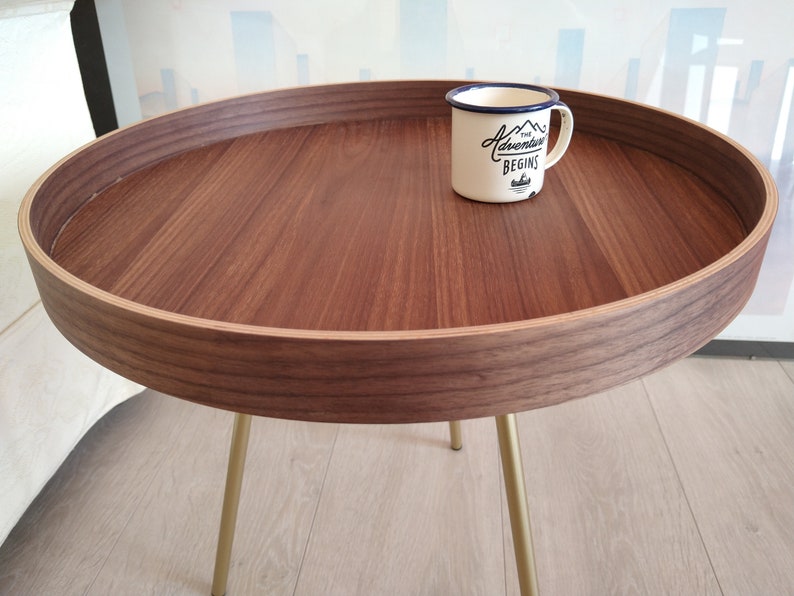 K&ouml;nnte beinhalten: Ein runder Holz-Beistelltisch mit einem goldenen Metallgestell. Die Tischplatte hat einen erh&ouml;hten Rand und eine helle Holzfurnier. Eine wei&szlig;e Emailletasse mit blauem Rand und dem Text "The Adventure Begins" steht auf dem Tisch.