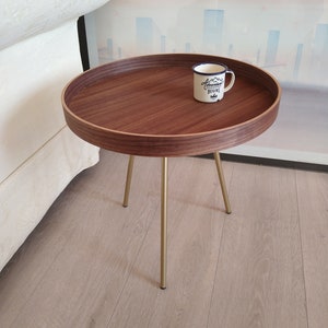 K&ouml;nnte beinhalten: Ein runder Holz-Beistelltisch mit goldenem Metallgestell. Der Tisch hat einen erh&ouml;hten Rand und eine wei&szlig;e Emailletasse mit dem Text "Adventure Begins" darauf.