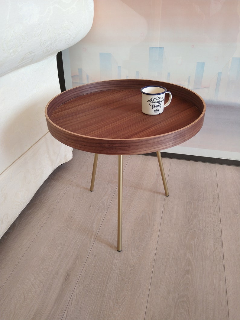 K&ouml;nnte beinhalten: Ein runder Holz-Beistelltisch mit goldenem Metallgestell. Der Tisch hat einen erh&ouml;hten Rand und eine wei&szlig;e Emailletasse mit dem Text "Adventure Begins" darauf.