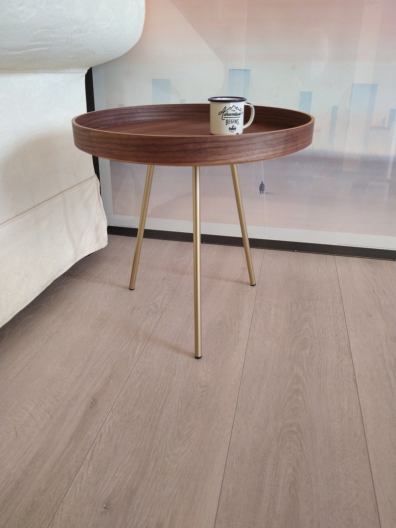 K&ouml;nnte beinhalten: Ein runder Holz-Beistelltisch mit goldenem Metallgestell. Der Tisch hat eine braune Holzplatte und drei goldene Beine. Eine wei&szlig;e Emailletasse mit schwarzem Text, der "Adventure Begins" lautet, steht auf dem Tisch.