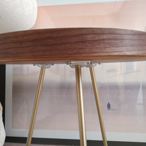 K&ouml;nnte beinhalten: Ein runder Holztisch mit Walnuss-Finish und drei goldenen Metallbeinen. Die Tischplatte ist glatt und hat eine leichte Rundung an der Kante.