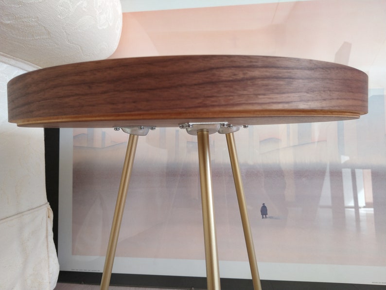 K&ouml;nnte beinhalten: Ein runder Holztisch mit Walnuss-Finish und drei goldenen Metallbeinen. Die Tischplatte ist glatt und hat eine leichte Rundung an der Kante.
