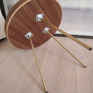 K&ouml;nnte beinhalten: Ein runder Holztisch mit einer nat&uuml;rlichen Holzoptik und drei goldenen Metallbeinen. Der Tisch steht auf einem hellen Holzboden.