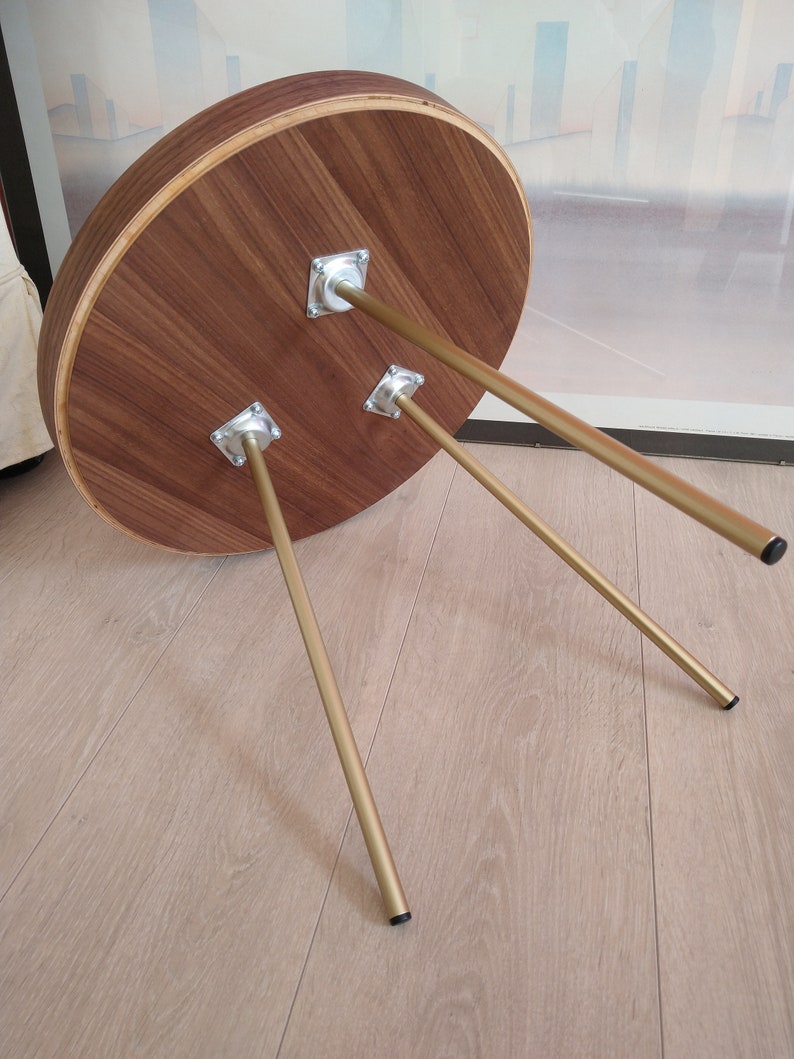 K&ouml;nnte beinhalten: Ein runder Holztisch mit einer nat&uuml;rlichen Holzoptik und drei goldenen Metallbeinen. Der Tisch steht auf einem hellen Holzboden.