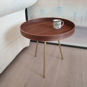 K&ouml;nnte beinhalten: Ein runder Holz-Beistelltisch mit Walnuss-Finish und goldenen Metallbeinen. Der Tisch hat eine wei&szlig;e Keramiktasse mit dem Text "Adventure Begins" darauf.