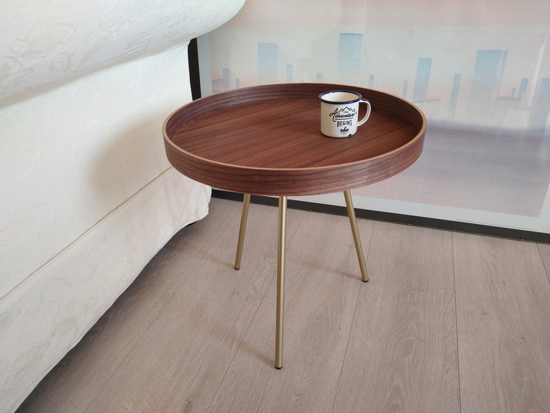 K&ouml;nnte beinhalten: Ein runder Holz-Beistelltisch mit Walnuss-Finish und goldenen Metallbeinen. Der Tisch hat eine wei&szlig;e Keramiktasse mit dem Text "Adventure Begins" darauf.