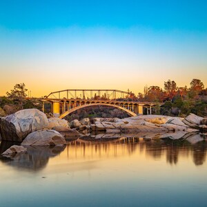 May include: A golden bridge arches over a calm river with large rocks in the foreground. The sky is a vibrant blue with a soft orange sunset in the background.