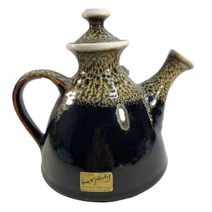 May include: A ceramic teapot with a glossy black base and a textured, mottled beige and brown glaze on the upper section and lid. It features a curved handle, a flared spout, and a gold-colored label reading Louis Mulcahy, Dingle, Ireland.