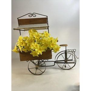 May include: A decorative metal bicycle cart with a wooden box filled with bright yellow flowers. The cart has a small wooden seat and a decorative metal canopy. The wheels are made of metal and the cart is set against a white background.