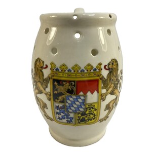 May include: Cream-colored ceramic container with a decorative design. The container features a coat of arms with two lions and a detailed crest. It has several small holes and a single hole at the top.