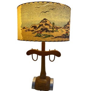 May include: Vintage table lamp with a desert landscape design on the lampshade. The shade features a tan background with a mountain range, cactus, and birds. The wooden base has a unique design with metal accents.