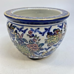 May include: A round ceramic planter with a blue rim and floral design. The planter features a white base with blue, gold, and pink floral patterns. The rim is trimmed with gold. The planter is suitable for small plants or as a decorative item.