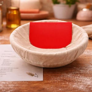 Op de afbeelding: Een beige rijsmand met een rode deegschraper erin, op een houten oppervlak. Een receptvel is zichtbaar. De scène suggereert bakken. De mand heeft een diameter van ongeveer 20 cm.