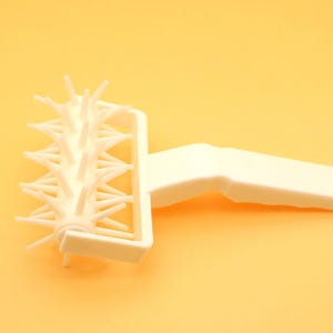 Può includere: Un bucatutto per pasta in plastica bianca, uno strumento da forno con un manico e un rullo ricoperto di piccole punte. Lo strumento è progettato per la cottura, su sfondo giallo.