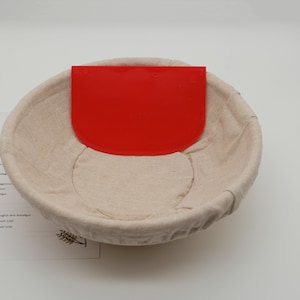 Może przedstawiać: Koszyk do wyrastania ciasta w kolorze beżowym z czerwoną plastikową szpatułką do ciasta w środku. Koszyk jest wykonany z plecionego materiału i ma okrągły kształt. Szpatułka ma kształt półkola i jest wykonana z plastiku.